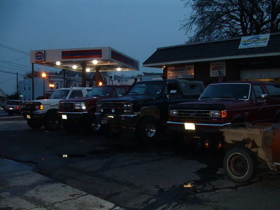 Several SUVs parked in a row at a gas station at dusk. One is a red Ford Bronco