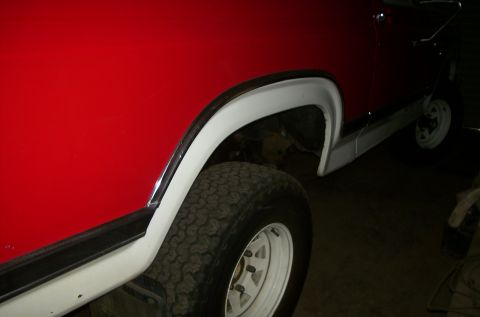 1981 Ford Bronco - The side of a red SUV is visible, with white fender flares and a white wheel.