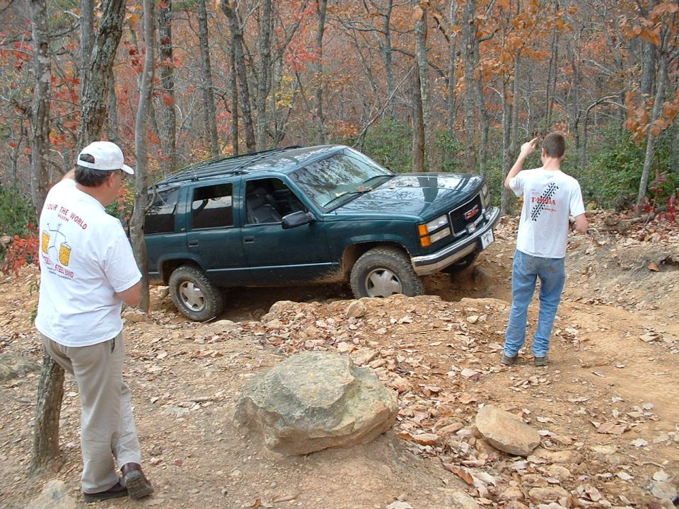 Green GMC Yukon climbing an off-camber rocky forest trail with two spotters