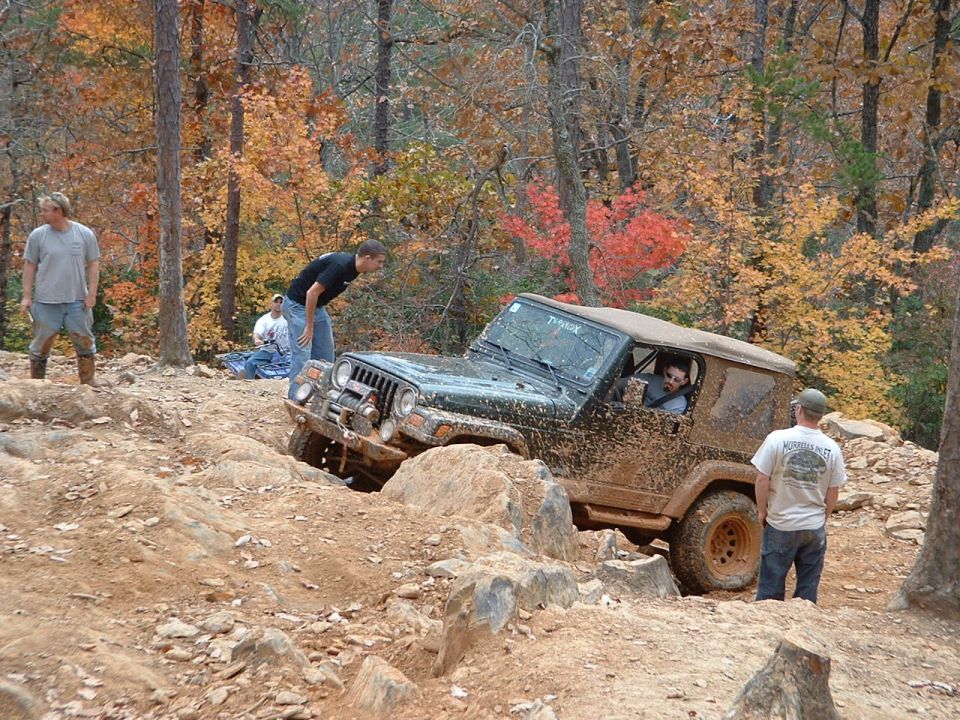 Muddy green Jeep Wrangler with tan soft top flexing over rocks while spotters watch