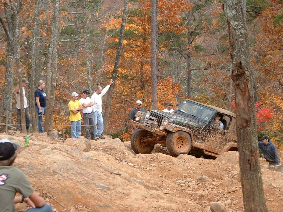 Mud-covered Jeep Wrangler navigating a rocky off-road trail with spectators watching