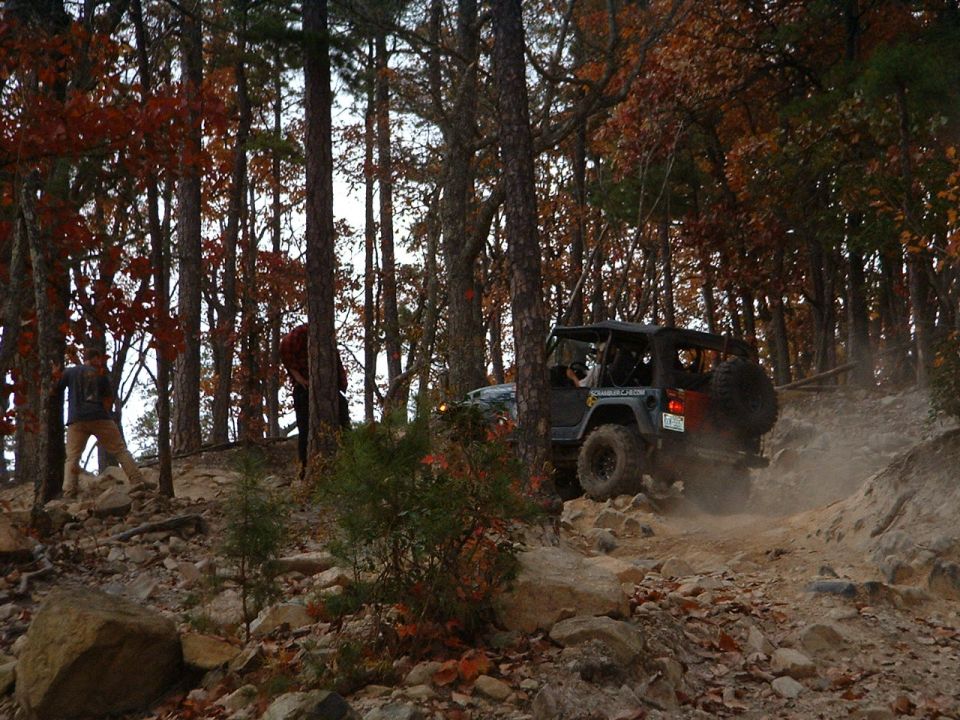 Gray soft-top Jeep Wrangler climbing a dusty rock trail in autumn woods
