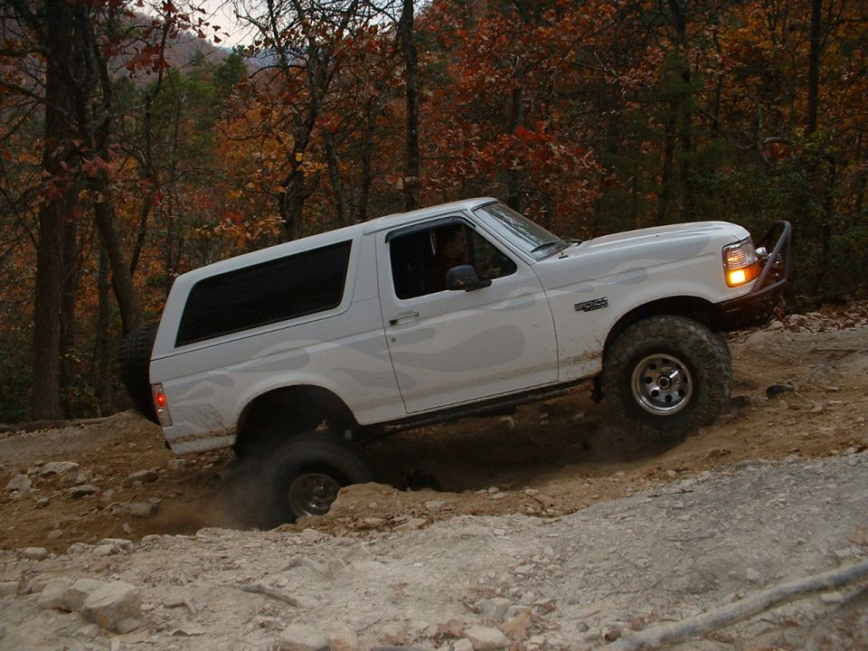 Lifted white Ford Bronco with ghost flames climbing a rocky hill, driver side view
