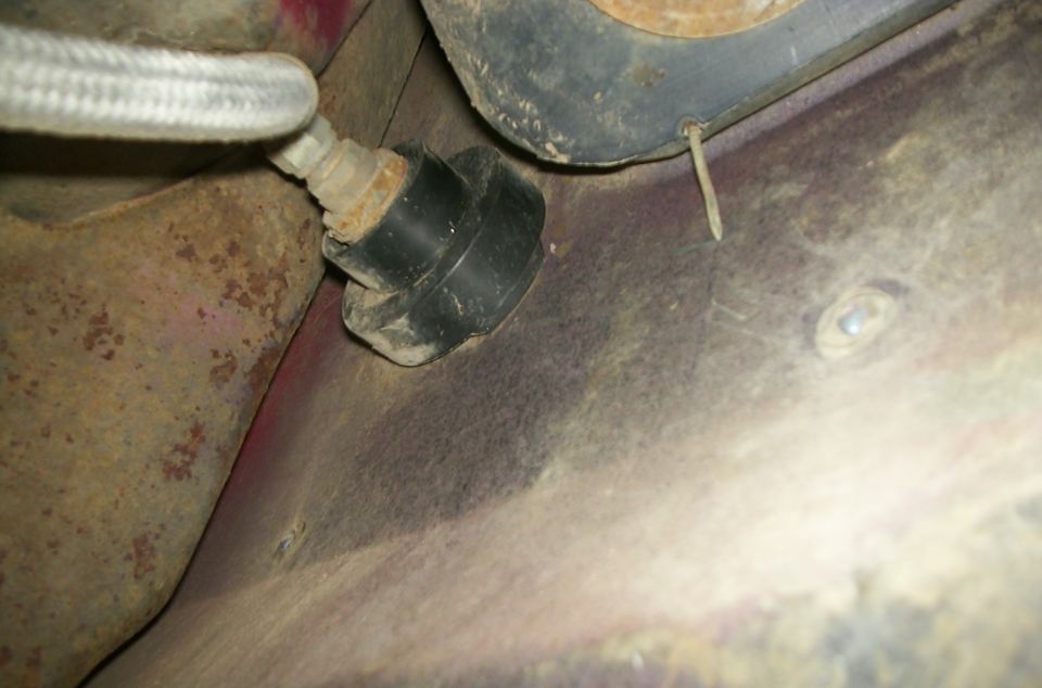 Underside of vehicle showing a black rubber grommet attached to rusty metal and a braided metal hose