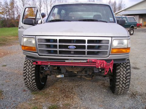 1996 Ford Bronco - A silver 1996 Ford Bronco is shown from the front, equipped with a red Hi-Lift jack mounted on its bumper. Another Bronco is visible in the…
