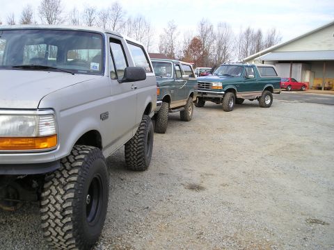 1996 Ford Bronco - A line of three Ford Broncos, including a silver one in the foreground and two green ones behind it, are parked on a gravel surface.