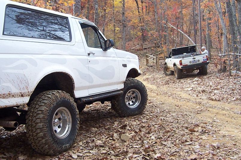 White lifted Ford Bronco on mud tires on a leaf-covered trail, second truck behind