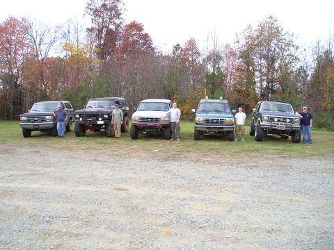 1996 Ford Bronco - A group of off-road vehicles are lined up with people standing next to them in a gravel lot.