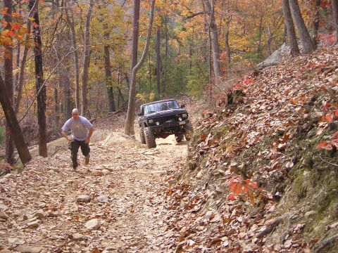 1996 Ford Bronco - A man walks on a rocky trail as a black SUV navigates the off-road terrain during autumn.