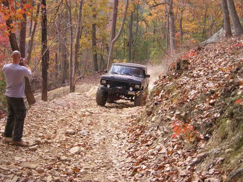 1996 Ford Bronco - A black 1996 Ford Bronco navigates a challenging off-road trail during the fall season, with a photographer capturing the action.