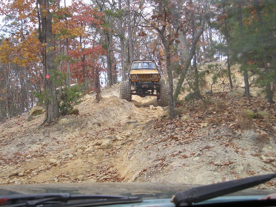 Yellow off-road vehicle with large tires climbing a steep, rocky trail in a wooded area