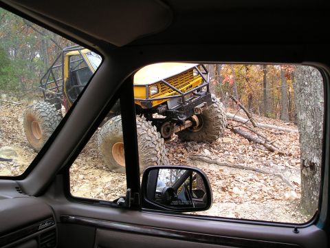 1996 Ford Bronco - A heavily modified yellow off-road vehicle navigates a challenging trail in a wooded area.