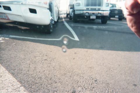 1990 Ford Bronco - washed-out parking lot scene framed between a white box truck and a white medium-duty cab-over truck, a fingertip intruding at the upper ri…