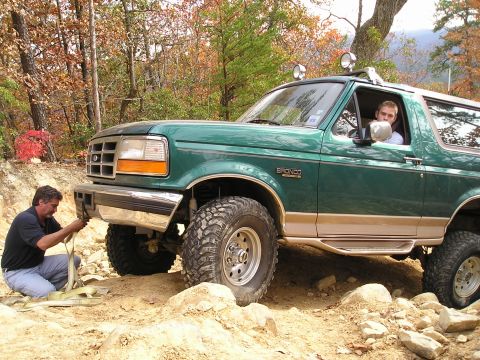 1996 Ford Bronco - A man secures a tow strap to the front of a green 1996 Ford Bronco as it navigates a challenging off-road course.