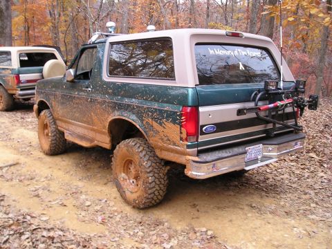 1996 Ford Bronco - Two muddy Ford Broncos are parked on a dirt road surrounded by autumn trees.