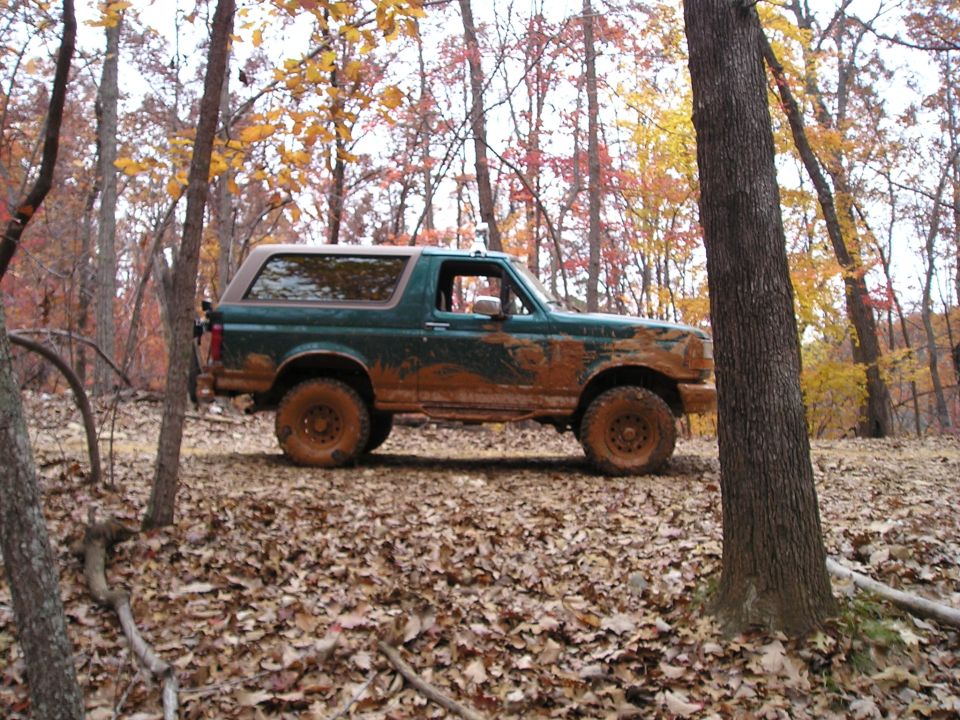 Mud-covered green 1992 Ford Bronco parked on a leaf-strewn forest floor