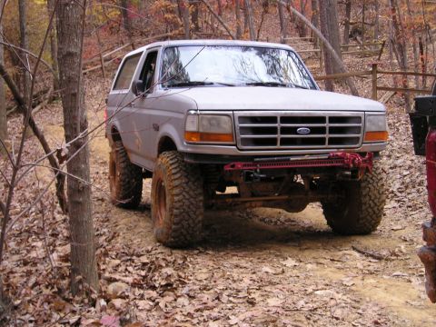 1996 Ford Bronco - A lifted 1996 Ford Bronco navigates a wooded trail during autumn, its large tires gripping the dirt path.
