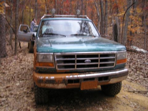 1996 Ford Bronco - A muddy green Ford truck is parked in a wooded area during autumn, with another vehicle visible behind it.