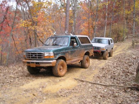 1996 Ford Bronco - Two Ford Broncos navigate a muddy trail during autumn, with colorful foliage in the background.