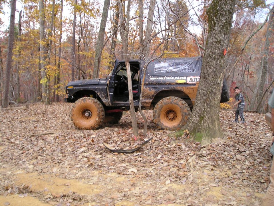 Muddy black 1978 Ford Bronco with large tires parked in a wooded area with fallen leaves
