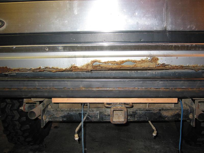 Silver SUV rear bumper with significant rust damage along the bottom edge, a black step bar, and a trailer hitch receiver. A wooden plank is temporarily placed above the hitch