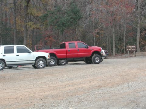 1985 Ford Bronco - A red six-wheeled pickup truck and a white SUV are parked side-by-side in a gravel lot near a wooded area.