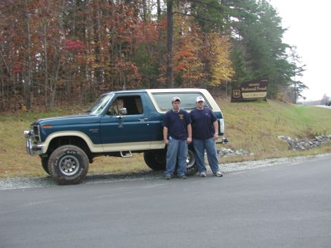 1985 Ford Bronco - A blue and tan Ford Bronco is parked on the side of a road with two men standing next to it, with a forest and a national forest sign in th…