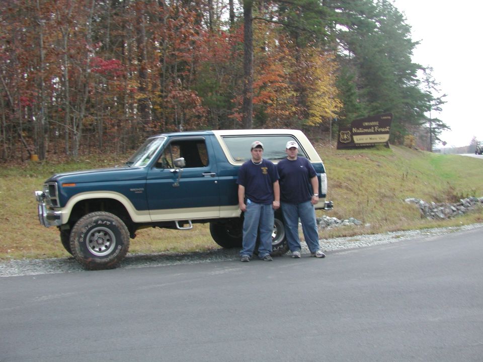 Blue and tan Ford Bronco with two men standing beside it, parked on a paved road near a forest