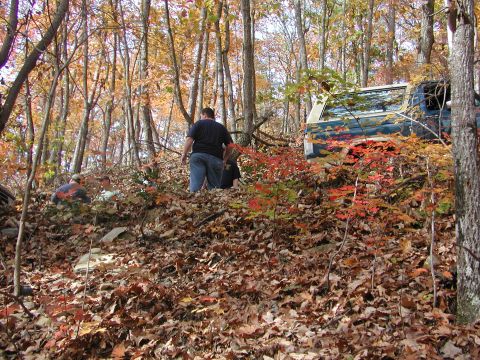 1985 Ford Bronco - A blue SUV is seen off-roading through a wooded area during autumn, with several people nearby.