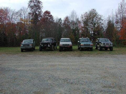 1985 Ford Bronco - Several SUVs are parked in a line on a gravel lot with trees in the background.