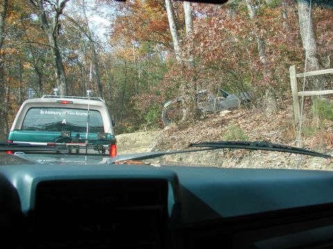 1985 Ford Bronco - SUV with 'In Memory of Tim Kramer' sticker drives on a wooded dirt road, with another vehicle visible off the side.