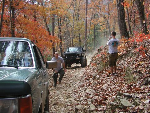 1985 Ford Bronco - Two trucks navigate a challenging off-road trail during autumn, with fall foliage visible in the surrounding trees.
