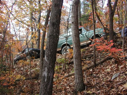 1985 Ford Bronco - Two off-road vehicles navigate a steep, wooded incline during autumn.