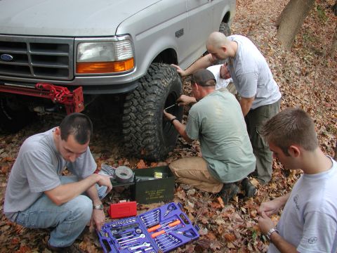 1985 Ford Bronco - Four men work on changing a tire on a silver truck in a wooded area with fallen leaves.