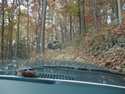 1985 Ford Bronco - Two off-road vehicles navigate a challenging rocky trail in a wooded area during autumn.