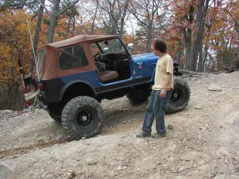 1985 Ford Bronco - A man stands next to a lifted Jeep CJ-7 on a rocky, wooded trail during autumn.