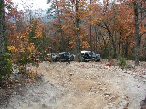 1985 Ford Bronco - Multiple off-road vehicles navigate a challenging, rocky path through a forest with autumn foliage.