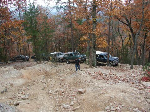 1985 Ford Bronco - Several off-road vehicles navigate a rocky trail in a wooded area during autumn.