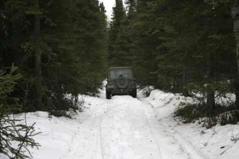 2003 Jeep TJ - A brown Jeep TJ drives on a snow-covered road through a dense forest.