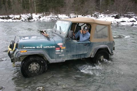 2003 Jeep TJ - A man drives a green Jeep Wrangler through a shallow river with snow-covered banks in the background.
