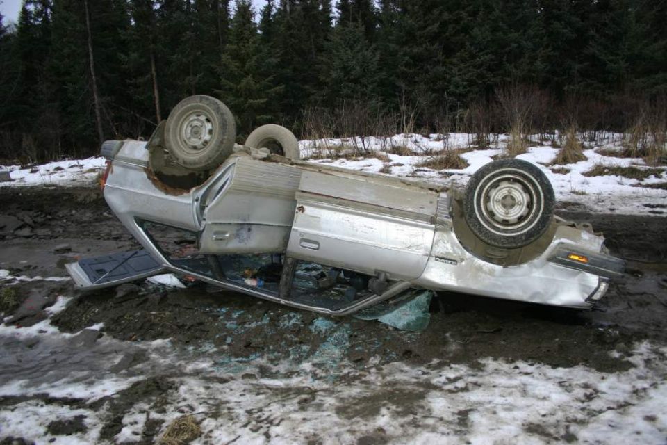 Overturned silver sedan in a muddy, snowy ditch with trees in the background