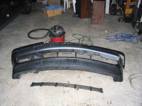 1995 Ford Taurus - A detached front bumper and grille assembly lies on a concrete floor, with a shop vacuum and other items in the background.