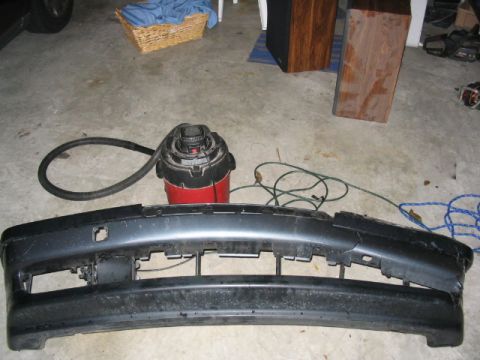 1995 Ford Taurus - A detached car bumper lies on a garage floor next to a shop vacuum and various cords.