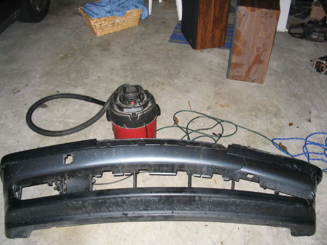 Car bumper and shop vac on concrete floor. Basket with blue cloth and speakers in background