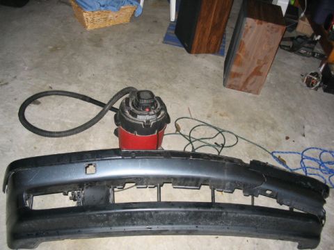 1995 Ford Taurus - A detached car bumper lies on a concrete floor next to a shop vacuum and various cords.