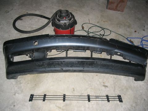 1995 Ford Taurus - A dark gray bumper and grille assembly are laid out on a concrete floor, with a shop vacuum visible behind them.