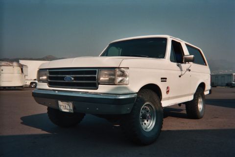 1990 Ford Bronco - white Ford Bronco full-size SUV on lifted suspension with large off-road tires, parked on open pavement with hills in the background.