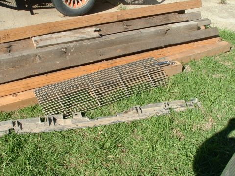1990 Ford Bronco - A chrome grille and a black plastic bumper part are laid out on the grass near some wooden planks.