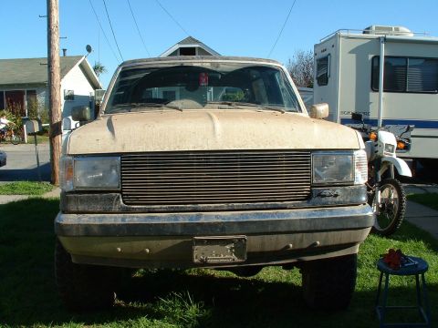 1990 Ford Bronco - The front grille of a tan Ford Bronco is visible, with an RV and a dirt bike parked behind it.