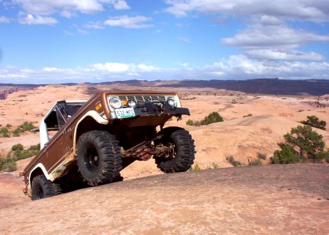 1974 Ford Bronco - A 1974 Ford Bronco navigates a rocky desert terrain under a cloudy blue sky.
